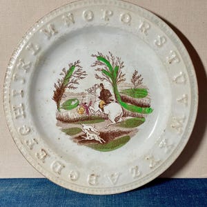 May include: A round white ceramic plate featuring an embossed alphabet border from A to Z. The center displays a colorful transfer print of a person riding a white horse, accompanied by two small dogs in a landscape with brown and green trees.