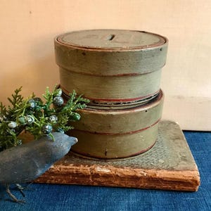 Puede incluir: Dos cajas redondas de madera apiladas con tapas. Las cajas están pintadas de verde claro con ribetes rojos. Las cajas están sobre un libro antiguo.