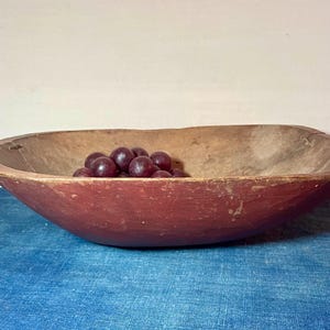 May include: A rustic, oval-shaped wooden bowl with a red exterior and a natural wood interior. The bowl contains a cluster of dark purple grapes. The bowl is sitting on a blue surface.
