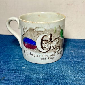 May include: Antique ceramic mug with a handle, featuring the letter "C" with a tiger stripe pattern, and the text "C begins at...and Cup." The mug has a cream-colored background with illustrations of a cat and other objects. The mug is on a blue surface.