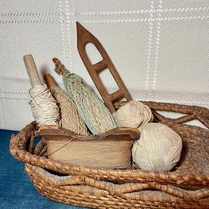 Puede incluir: Una cesta de mimbre llena de herramientas de tejido vintage. La cesta contiene carretes de madera, hilo y una lanzadera de madera. El hilo es de varios colores, incluyendo beige, azul claro y tonos naturales. La cesta está hecha de material marrón tejido.