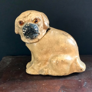 May include: A vintage, cream-coloured, papier-mâché dog figurine with black painted features and orange glass eyes. The dog is sitting with its head tilted to the side.