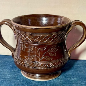 May include: A brown ceramic bowl with two handles, featuring etched floral and geometric patterns. The bowl has a wide opening and sits on a small, circular base. The color is a rich, dark brown.
