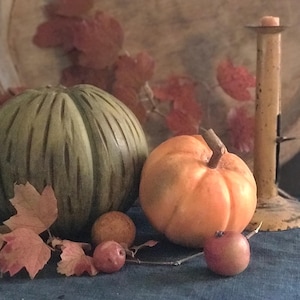 Puede incluir: Un bodegón con una calabaza verde y una calabaza naranja. Pequeñas manzanas rojas y esferas naranjas están dispuestas sobre una superficie azul. Hojas rojas y un candelabro completan la exhibición otoñal.