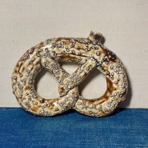 May include: A ceramic pretzel figurine with a glossy finish. The pretzel is brown with white and tan speckles, and has a small loop at the top. The figurine is on a blue surface.