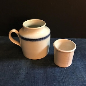 Może przedstawiać: Dwa białe ceramiczne kubki z niebieskimi ozdobami. Większy kubek ma uchwyt i niebieską wstążkę wokół górnej krawędzi. Mniejszy kubek jest krótszy i nie ma uchwytu.