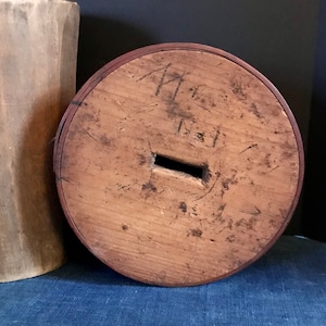 Puede incluir: Una tapa redonda de madera con un pequeño agujero rectangular en el centro. La tapa está marcada con números y letras escritas a mano, incluyendo "1811".