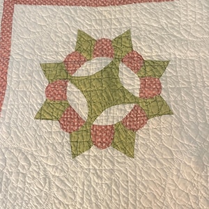 May include: A white quilted fabric featuring a central star-shaped applique pattern in sage green and a pink floral print. The quilt has a textured, stitched surface with a pink floral border and a sage green accent line along the top edge.