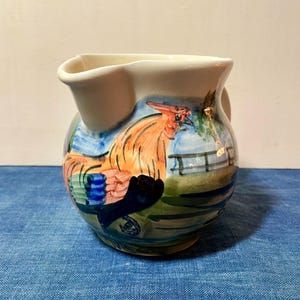 Puede incluir: Una jarra de cerámica con un diseño de gallo. La jarra presenta un borde y un asa blancos, con un gallo pintado a mano en naranja, azul y verde. El fondo incluye una valla y un cielo azul.