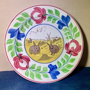 Puede incluir: Un plato redondo de cerámica blanca con una ilustración central de tres conejos marrones en un campo amarillo. El borde está decorado con un patrón repetitivo de flores rosas y azules unidas por enredaderas de hojas verdes sobre fondo blanco.