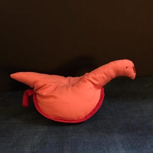 Puede incluir: Un pájaro de tela rosa con un ribete rojo alrededor de los bordes. El pájaro está acostado de lado con la cabeza girada hacia la derecha.