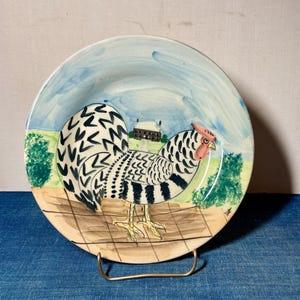May include: A round ceramic plate featuring a hand-painted illustration of a black and white speckled rooster standing on a brown grid-patterned ground. The background depicts a light blue sky, green bushes, and a small house in the distance.