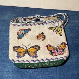 May include: A rectangular beaded pouch featuring a cream-colored background with multiple butterfly designs in blue, yellow, orange, and pink. The bag includes a green bottom border with the text Jane M. Wightman and thin silver ribbon straps.