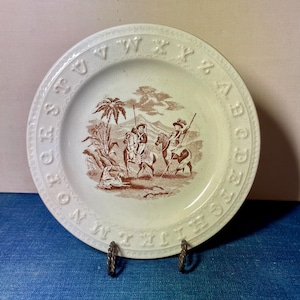 May include: A white ceramic plate with a brown transferware design. The plate features a scene of figures on horseback and a border with embossed letters. The plate is displayed on a metal stand.