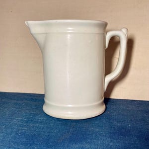 May include: A solid off-white ceramic pitcher featuring a cylindrical body, a flared base, a prominent pouring spout, and a curved handle attached to the side. The surface has a smooth, uniform finish with subtle horizontal ridges near the top and bottom.