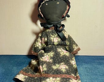 May include: Handmade cloth doll with a black face and a black bonnet trimmed with pink ribbon. The doll wears a dark floral print dress with pink accents and a black bow at the neck. The doll's feet are clad in white socks.