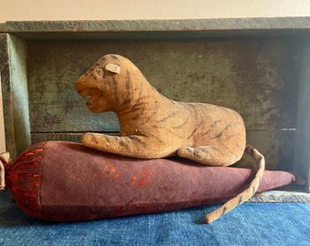 Puede incluir: Un juguete de peluche de tigre rayado marrón y negro acostado sobre una almohada de terciopelo burdeos. El tigre está acostado de lado con la cabeza girada hacia la izquierda. La almohada está en una caja de madera con un interior pintado de verde.