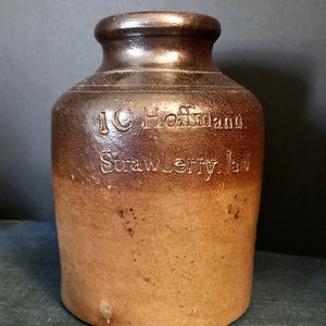 Pode incluir: Um pote de cerâmica marrom com uma boca larga e um acabamento envidraçado. O pote está etiquetado com "J.C. Hoffman Strawberry Jam".