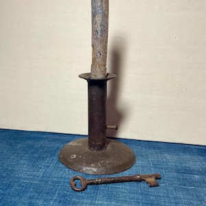 May include: An antique metal candlestick with a lit candle and a vintage key. The candlestick is dark brown with a round base and a tall, cylindrical stem. The candle is a light brown color. The key is also dark brown.