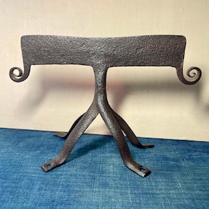 May include: A dark brown, hand-forged metal stand with a textured surface. The stand has a rectangular top with curled ends and three legs. The legs are joined at the center and splay outward.