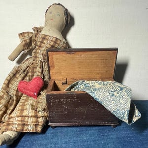 May include: A vintage cloth doll wearing a brown and tan plaid dress with a small red heart attached to the front, positioned next to an open dark brown wooden chest containing a blue and white patterned fabric piece with a floral design.