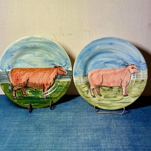 May include: Two round ceramic plates featuring hand-painted illustrations of cows standing in a green field under a blue sky. The left plate shows a brown cow, while the right plate depicts a light pink cow. Both plates have a glossy finish.