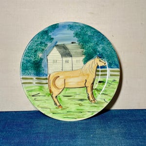 May include: A round ceramic decorative plate featuring a hand-painted scene of a light tan horse standing in a green pasture. In the background, there is a white building with a dark roof and a white fence, all surrounded by lush green trees.