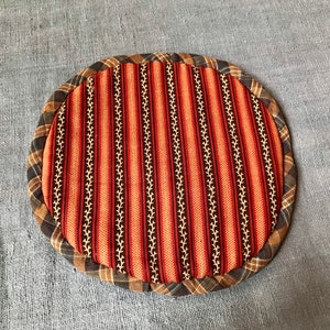 Puede incluir: Un posavasos redondo de tela a rayas naranja y marrón con un patrón floral blanco y negro. El posavasos está hecho de una tela suave acolchada y tiene un borde con flecos.