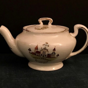 Pode incluir: Bule de chá de cerâmica branca com acabamento dourado e uma cena pintada de uma igreja e casas. A chaleira tem uma tampa com uma pega no topo. O texto na chaleira diz "Church Street, Park Hill, The House, And Green Walls".