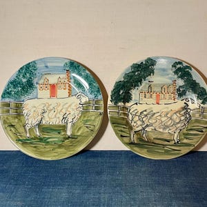 May include: Two round ceramic plates featuring hand-painted illustrations of a white sheep standing in a green pasture. Each plate depicts a house with a red door in the background, a wooden fence, and green trees, rendered in a loose, artistic style.