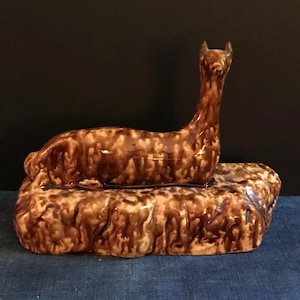 May include: A brown ceramic figurine of a llama lying down on a rock. The figurine is glazed in a brown and white mottled pattern.