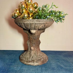 May include: A decorative, weathered-look pedestal planter with a tree trunk base. The planter holds a cluster of gold ornaments and green sprigs with blue berries. The planter is made of a dark, textured material.