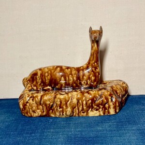 May include: A ceramic figurine of a deer in a resting position, featuring a glossy brown and cream drip glaze finish. The deer is depicted with an elongated neck and upright head, resting on a textured, rock-like base with matching glaze.