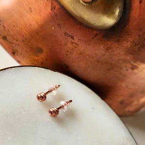 May include: A pair of small, rose gold stud earrings with a simple, coiled design. The earrings are displayed on a white surface.