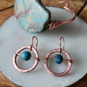 May include: A set of copper earrings with turquoise stones. The earrings are in the shape of a circle with a smaller circle in the center. The stones are held in place by a copper wire.