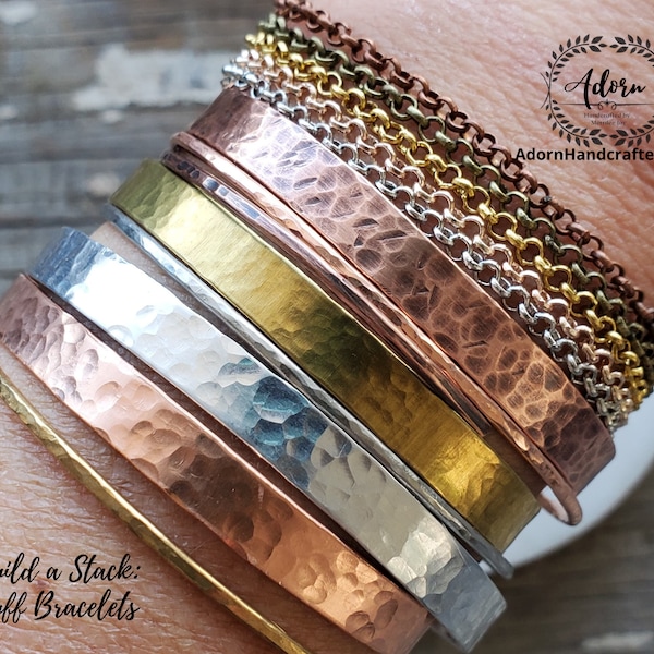 May include: A stack of hammered metal cuff bracelets in various colors, including copper, silver, and gold. The bracelets are arranged in a graduated size, with the largest at the bottom and the smallest at the top. The text "Build a Stack: Cuff Bracelets" is visible at the bottom of the image.