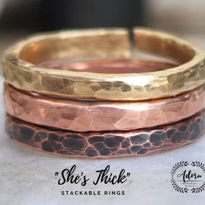 Può includere: Tre anelli impilabili in oro, rame e metallo nero. Gli anelli hanno una texture martellata. Il testo "She's Thick" è scritto sotto gli anelli. Il testo "Stackable Rings" è scritto sotto il testo "She's Thick".