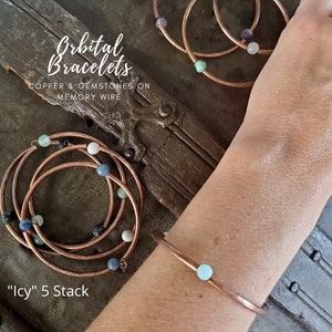Könnte beinhalten: Fünf Kupferdrahtarmbänder, die auf einem Handgelenk gestapelt sind. Jedes Armband hat eine einzelne Edelsteinperle in Blautönen, Grün und Weiß. Die Armbänder sind mit "Icy" 5 Stack beschriftet. Der Text "Orbital Bracelets" ist über den Armbändern sichtbar. Der Text "Copper & Gemstones on Memory Wire" ist ebenfalls sichtbar.