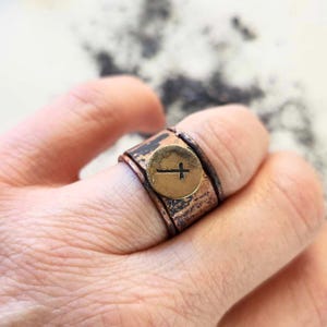 Può includere: Un anello in rame con finitura martellata e un ciondolo a croce in ottone. L'anello è indossato su un dito.