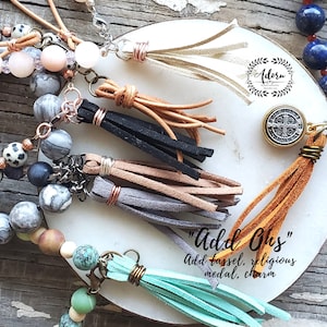 May include: A close-up of a variety of colorful tassels, beads, and charms. The tassels are made of leather and come in a variety of colors, including beige, brown, black, gray, and turquoise. The beads are made of various materials, including stone and glass. The charms include a religious medal and a letter "I". The text "Add Ons" is visible in the image, along with the text "Add tassel, religious medal, charm".