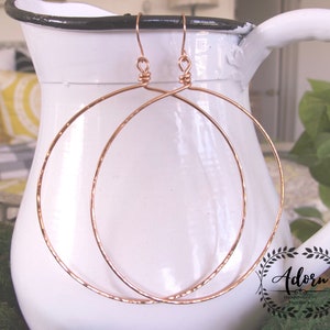 May include: A pair of large, hammered, rose gold hoop earrings. The earrings are made of wire and have a simple, elegant design.