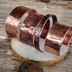 May include: Four copper bracelets with hammered textures and varying widths are displayed on a white and gold coaster. The bracelets have a warm, reddish-brown hue. The bracelets are approximately 1 to 2 inches wide.