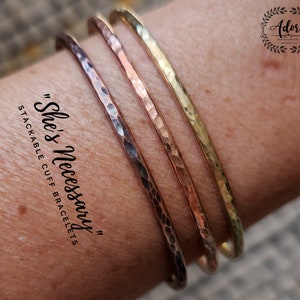 May include: Three hammered metal cuff bracelets in gold, copper, and silver tones. The bracelets are stacked on a person's wrist. The text "She's Necessary" and "Stackable Cuff Bracelets" is written on the image.