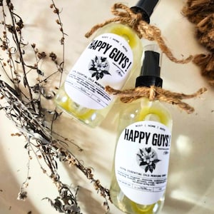 May include: Two clear glass bottles with yellow liquid and white labels. The labels have black text that reads "HAPPY GUYS!" and "100% ESSENTIAL OILS PERFUME SPRAY". The bottles are tied with twine and have a spray nozzle.