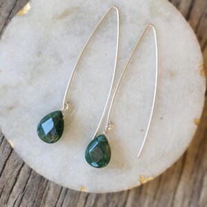 May include: A pair of silver dangle earrings with green gemstone drops. The earrings are on a white marble surface.