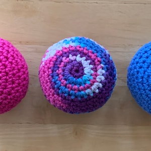 May include: Three crocheted balls, one pink, one purple and blue, and one blue. The balls are all the same size and are arranged in a row.