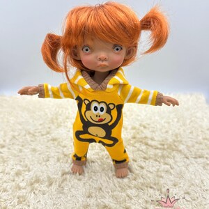 May include: A small doll with red hair wearing a yellow and white striped hooded jumpsuit with a monkey graphic. The doll is standing on a white fluffy rug.