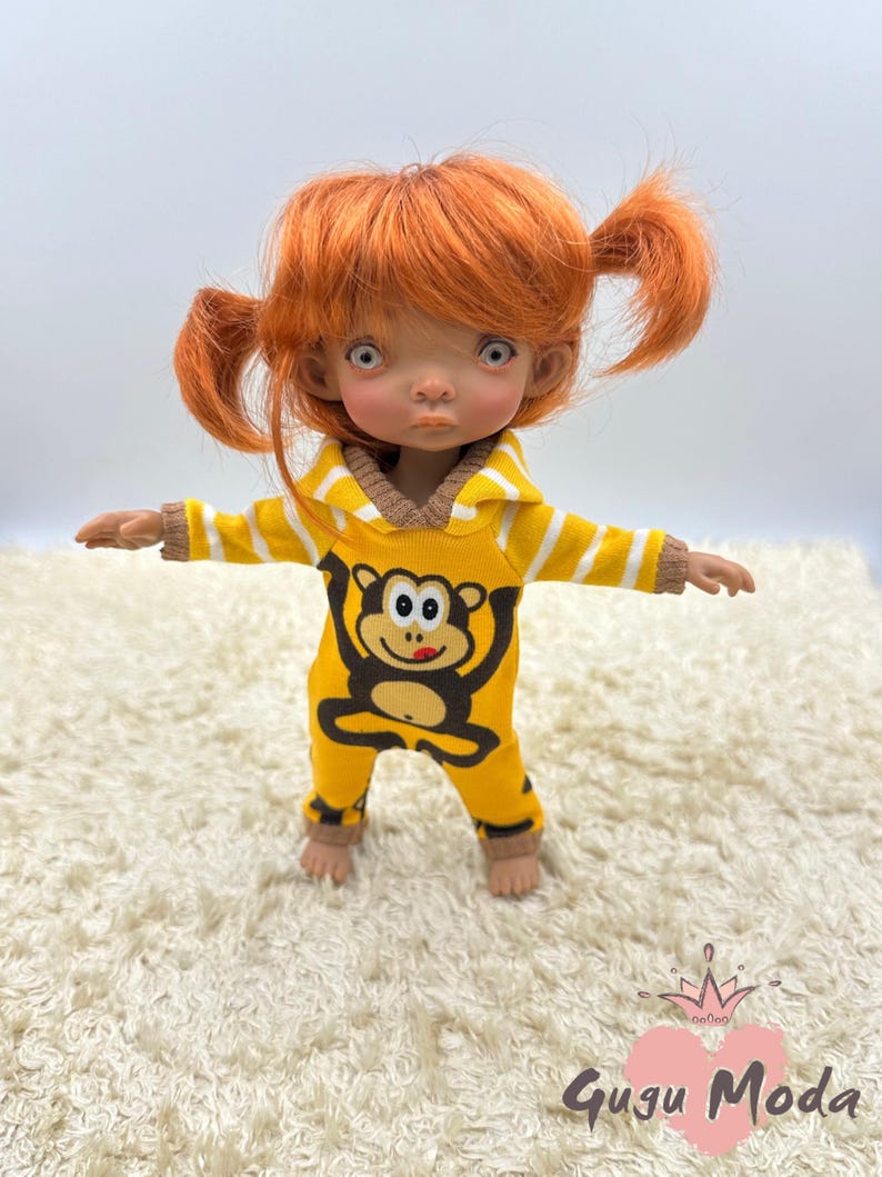 May include: A small doll with red hair wearing a yellow and white striped hooded jumpsuit with a monkey graphic. The doll is standing on a white fluffy rug.