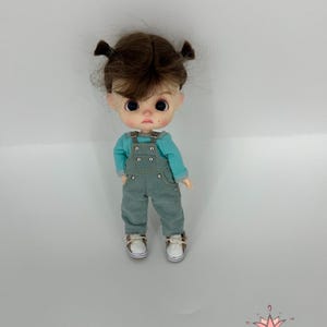 May include: A small doll with brown hair in pigtails wearing a blue and white outfit and white shoes. The doll is standing on a white background.