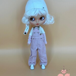 May include: A doll wearing a white corduroy hat, white wig, and pink overalls. The doll has large brown eyes, a white shirt with pink striped sleeves, and white sneakers. The doll is standing against a light brown background. The doll is wearing a white corduroy hat.